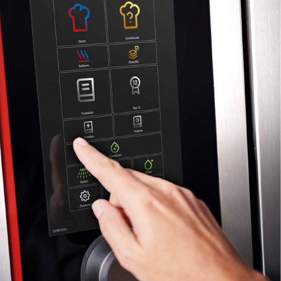 Detailní záběr prstu, který ovládá dotykové menu s různými ikonami funkcí jako vaření, CombiGuide, Retherm, MenuPlus, protokoly, nastavení a čištění.