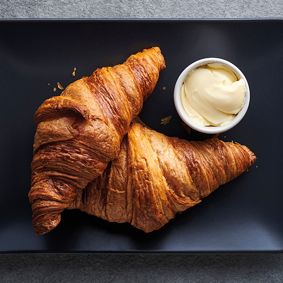 Dva zlatavé, čerstvě upečené croissanty na černém talíři s miskou másla – připravené pomocí CHEF'S COMBI od HOBARTU pro dokonalou texturu a zhnědnutí.