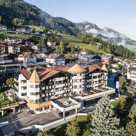 Letecký pohled na Gardena Grödnerhof v Ortisei, pětihvězdičkový hotel Relais & Châteaux s více než stoletou tradicí, zasazený do alpské krajiny údolí Gardena mezi zelenými svahy a tradičními domy.