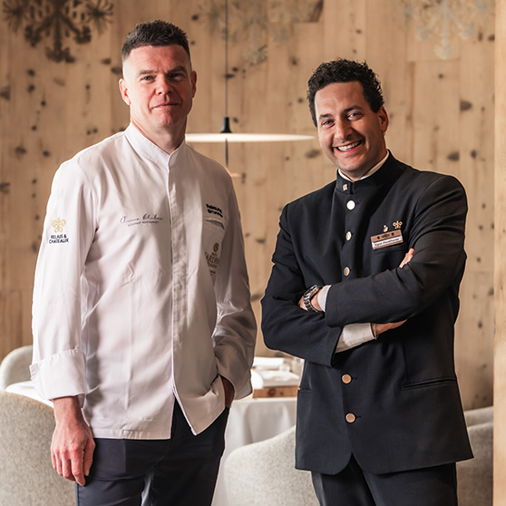 Dvě osoby ve formálním oblečení typickém pro hotelnictví stojí v interiéru restaurace s dřevěnými stěnami a připravenými stoly, v souladu s textem zdůrazňujícím důležitost bezpečných procesů a spolehlivé mycí technologie, jako jsou systémy HOBART.