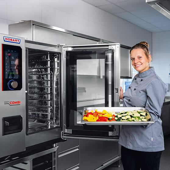 Profesionální combi steamer HOBART v moderní nerezové kuchyni, otevřené dveře, tác s čerstvou zeleninou jako paprika a cuketa připravený pro efektivní vaření v gastronomii