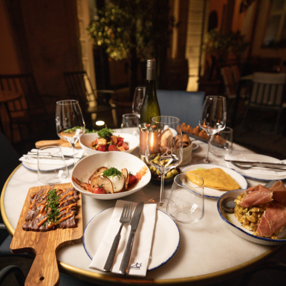 Venkovní restauranční stůl upravený s několika jídly, předkrmy, pečivem, lahví vína a různými sklenicemi.