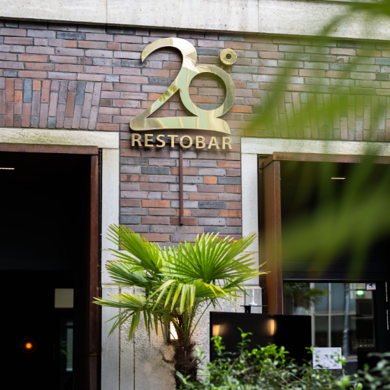 Vnější pohled na cihlovou budovu se zlatým nápisem 20° RESTOBAR nad vstupem, doplněný zelenými rostlinami.