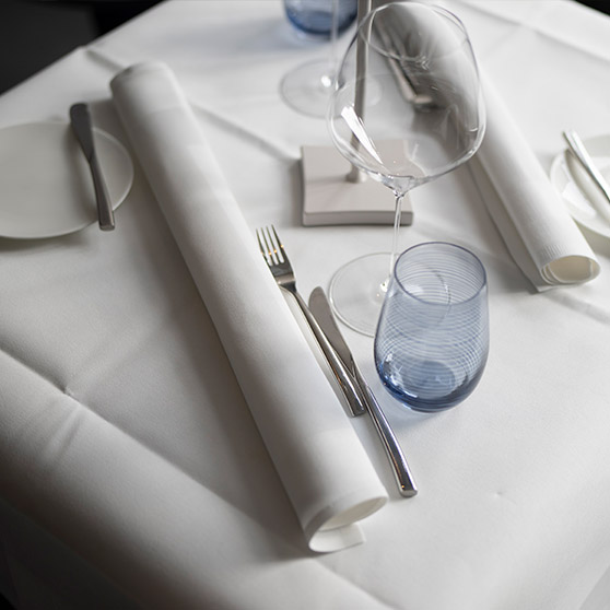 Elegantně prostřený stůl v restauraci s úhledně rozmístěným příborem, sklenkami na víno, srolovanými ubrousky a modrou sklenicí, který vytváří vytříbenou atmosféru.