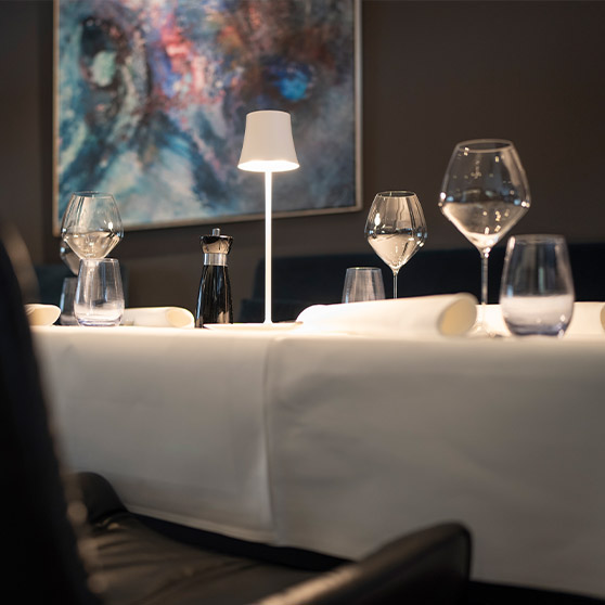 Detail elegantně prostřeného stolu v restauraci RIVA s kvalitní sklenicí, bílým ubrusem a moderním osvětlením.